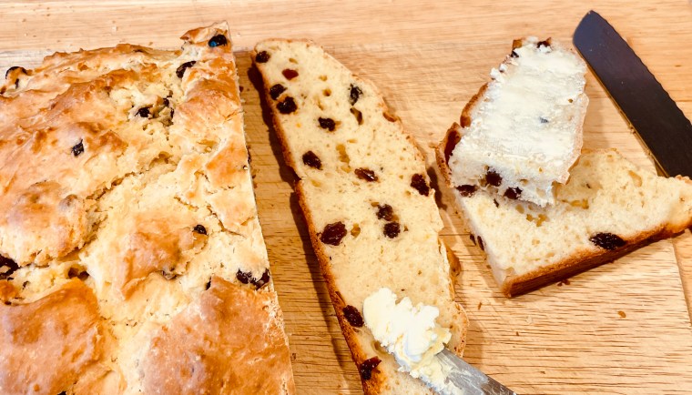 Slices of Irish soda bread, some with butter and some without are on a wooden cutting board. The loaf of soda bread is on the right side of the picture. A serrated knife and a butter knife are also featured.