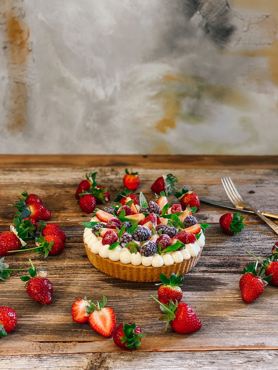 A berry pastry sits on an oak table with a cement background. Sliced and whole strawberries are scattered around the dessert. A fork and knife are also featured.