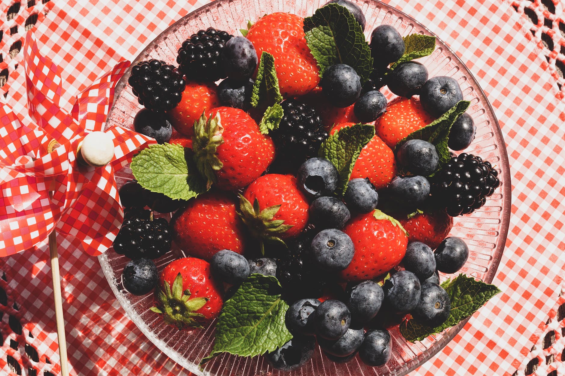 A glass bowl rests on a white and red checkered tablecloth. Inside the bowl are handfuls of blueberries, strawberries, and blackberries. The viewer sees the bowl and berries from overhead.