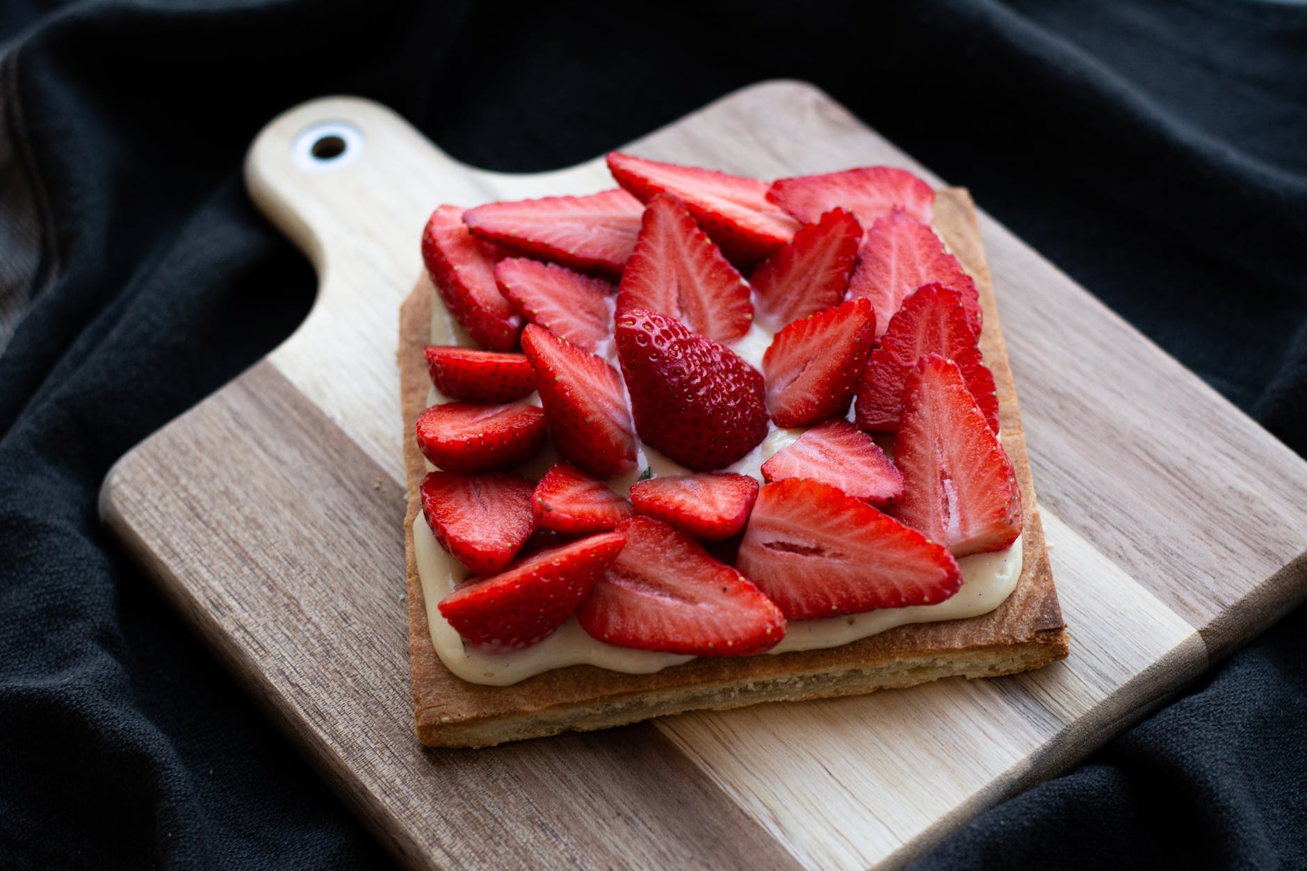 Sliced strawberries sit artfully on a piece of pastry. The food item sits on a wooden cutting board. The background is a piece of black fabric. The image is viewed slightly higher than eye level.